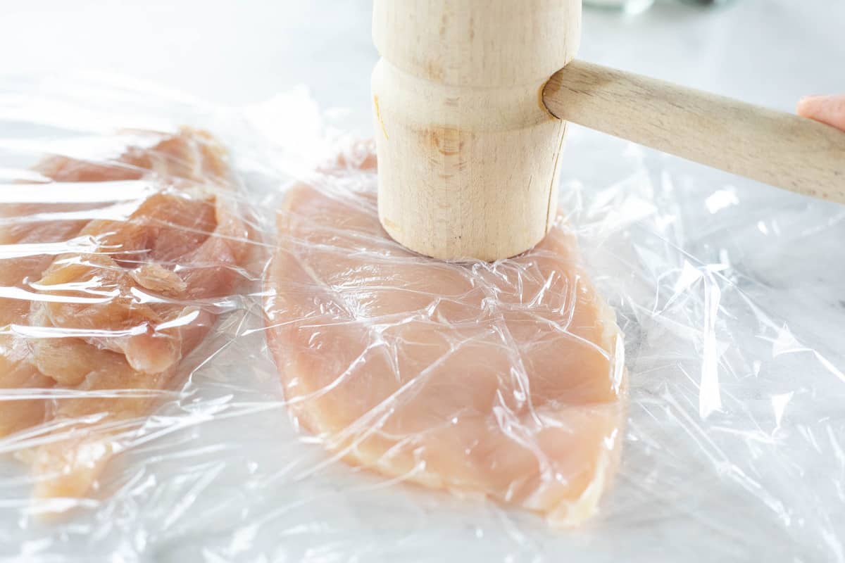 A kitchen mallet pounding thin sliced chicken breasts between two pieces of plastic wrap. 