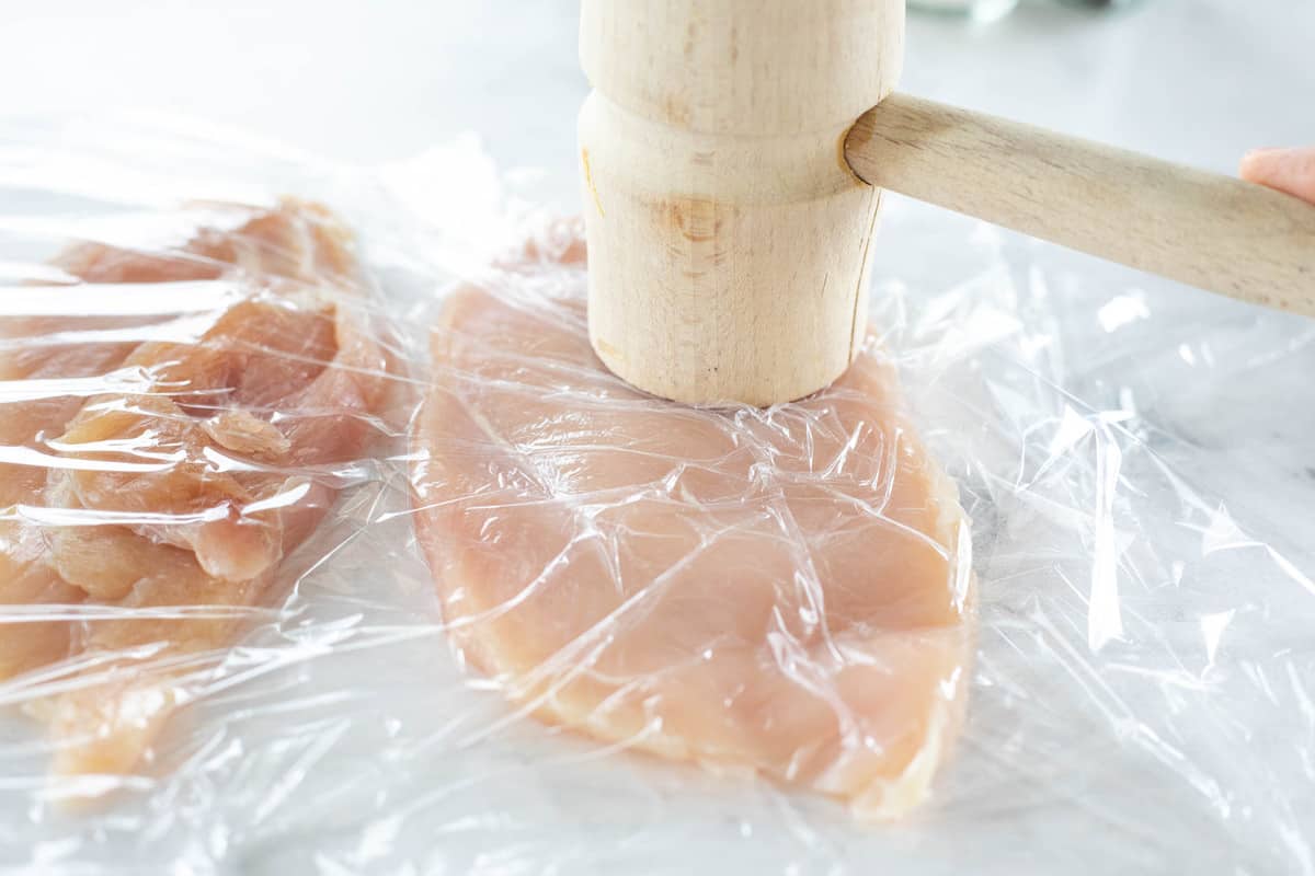 Uma cozinha martelo batendo fatias finas de peitos de frango entre dois pedaços de filme plástico. 