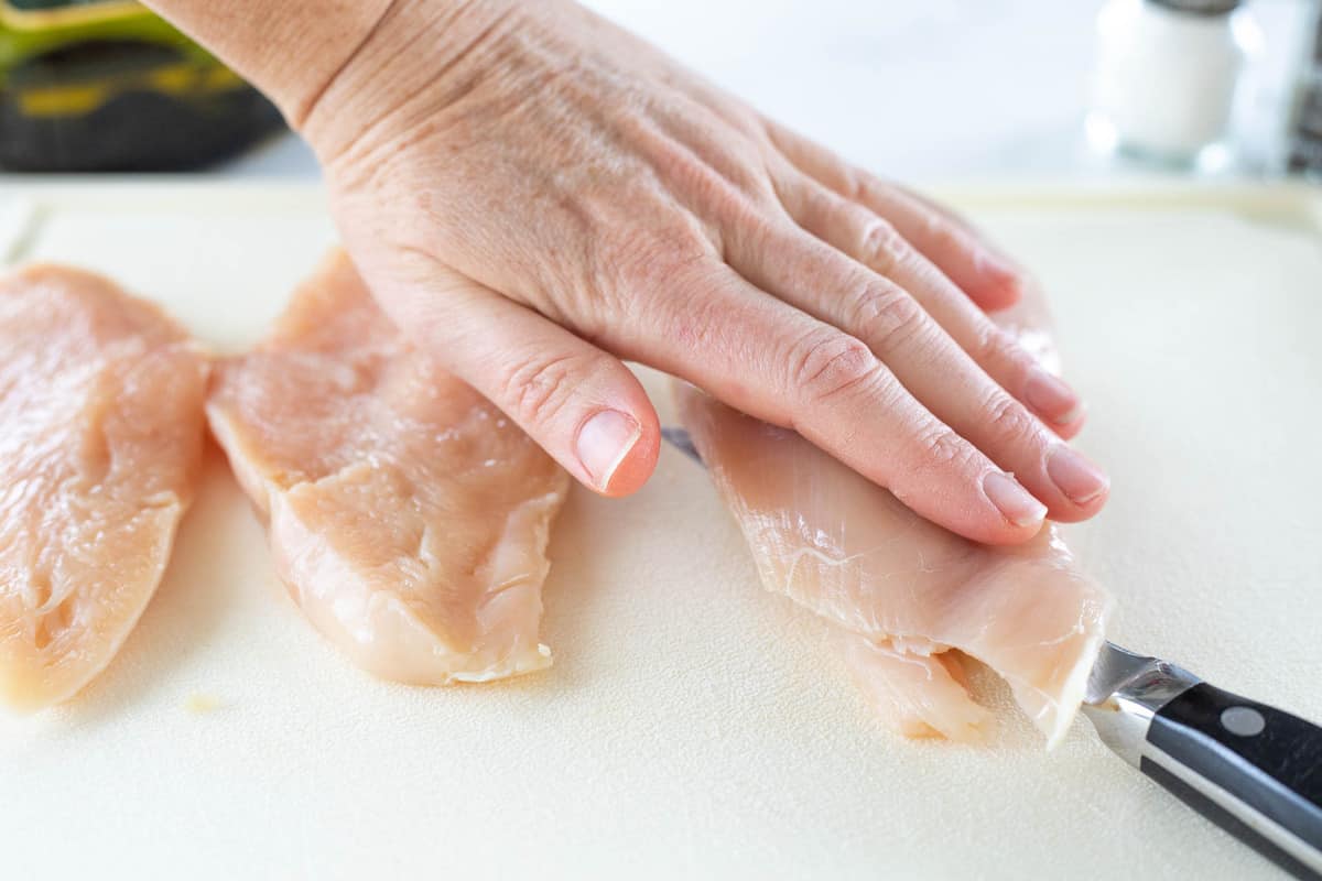 Chicken breasts shown being sliced in half on top of a cutting board with a knife. 