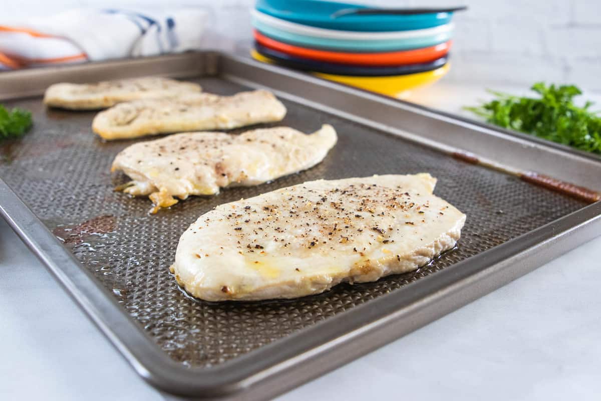 Seasoned thin sliced chicken breasts being shown on top of a baking sheet with a stack of plates behind the baking sheet.
