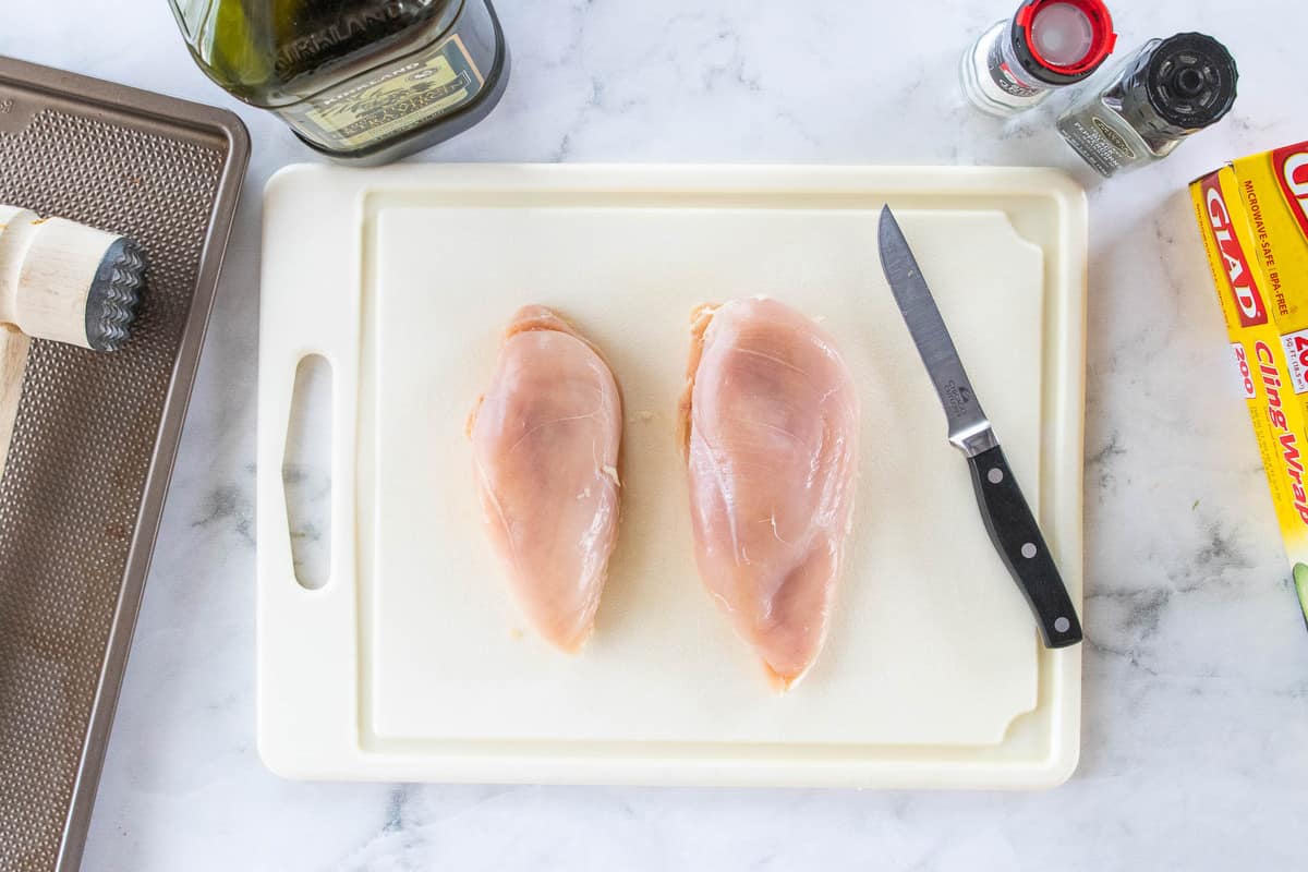 2 raw chicken breasts on a cutting board next to a knife. 
