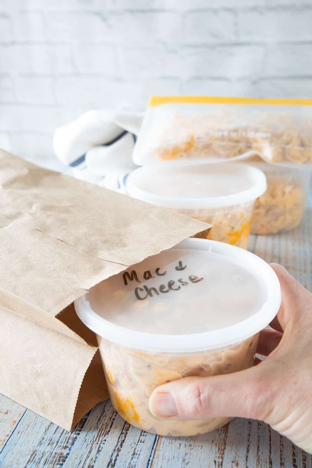 Cooked Mac & cheese in a container being placed by a hand into a brown paper bag. 