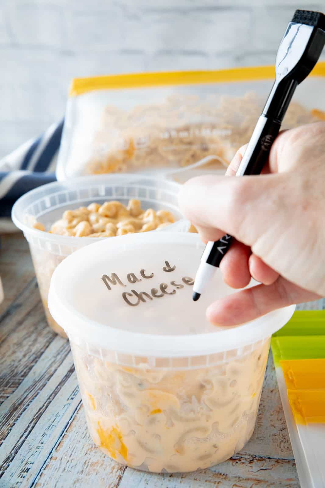 A hand writing Mac & cheese on top of a plastic lid that is on top of a plastic container filled with pasta.