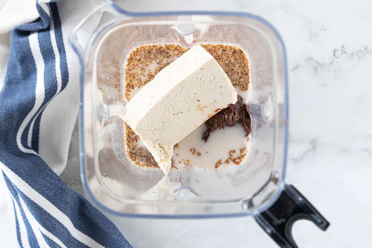 Milk, flax seed, Nutella and tofu being shown in a blender that is on top of a granite countertop with a blue dish towel next to the blender. 