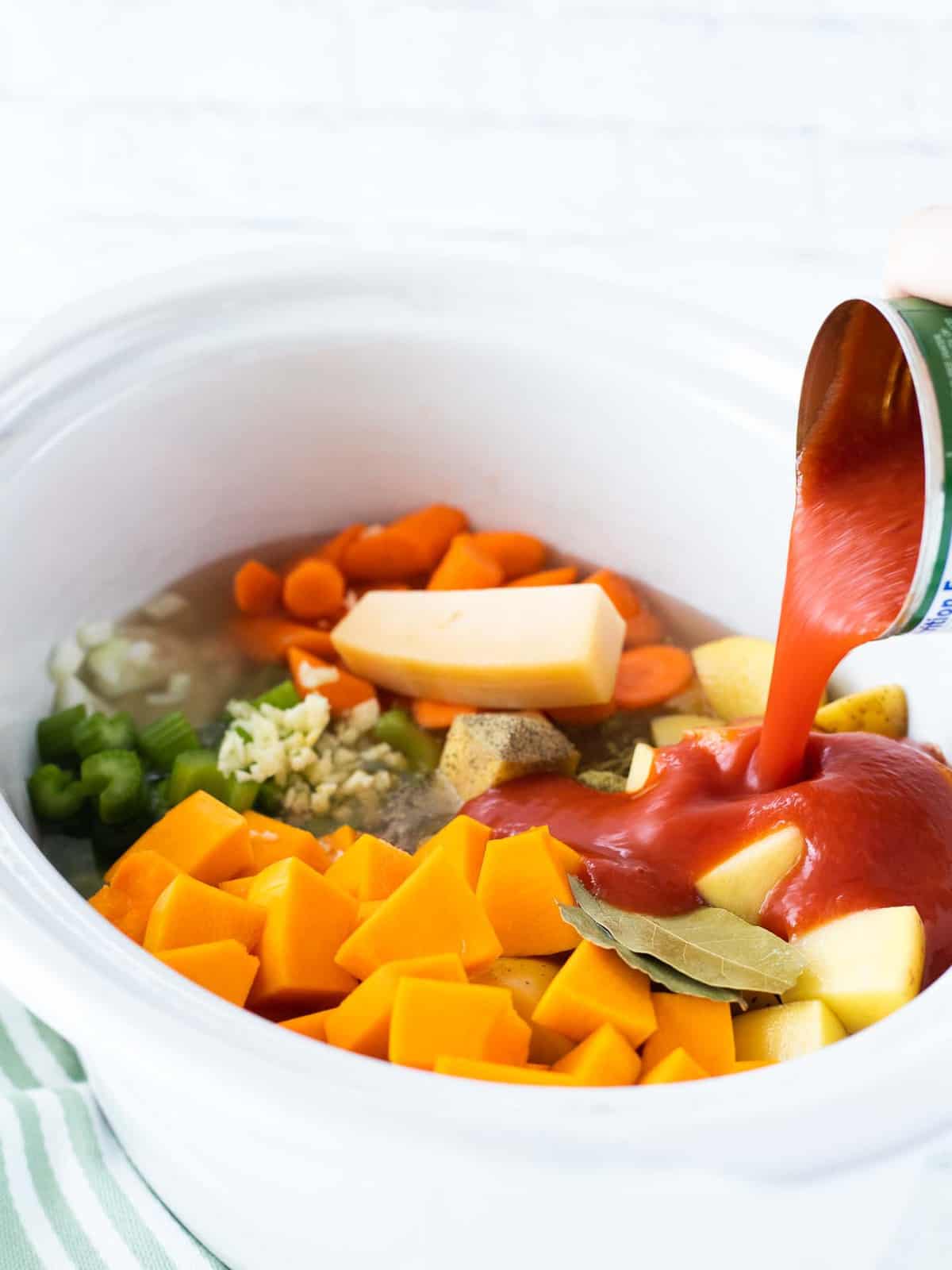 A can of tomato sauce being poured over chunky vegetables in a crockpot.