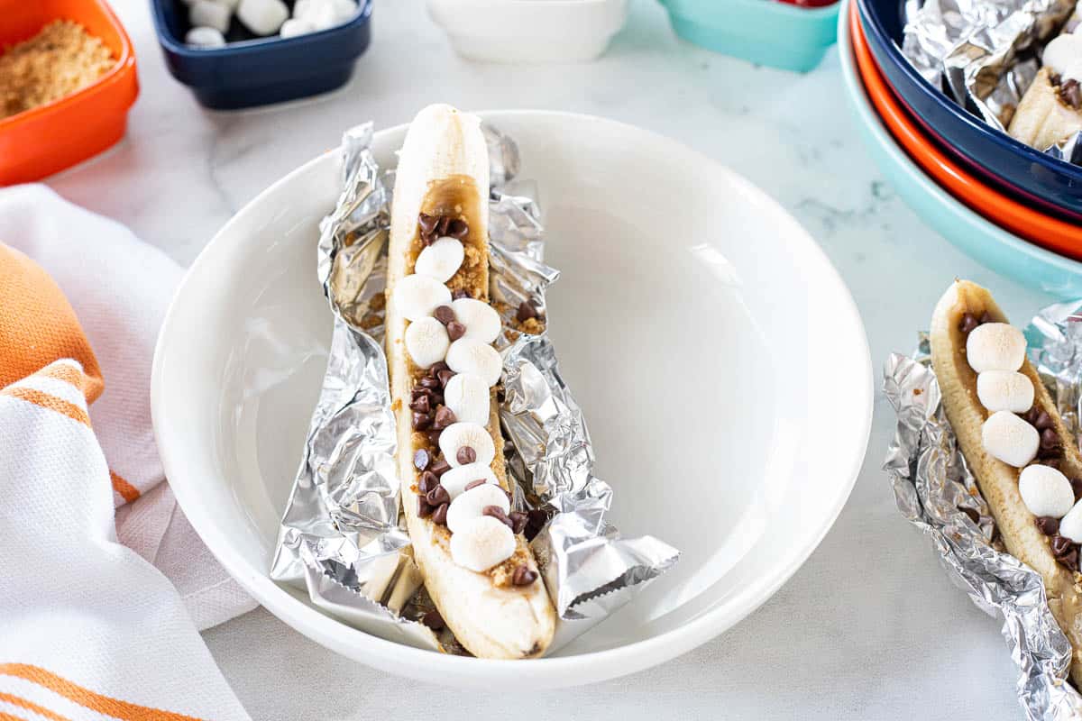 A S'mores banana boat wrapped in tin foil being shown in a white bowl.
