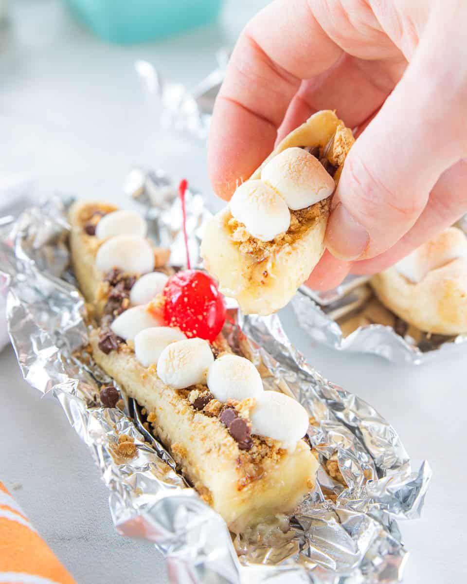 A hand grabbing a bite of a tasty dessert being shown with the rest of the s'mores banana boat dessert wrapped in tin foil.