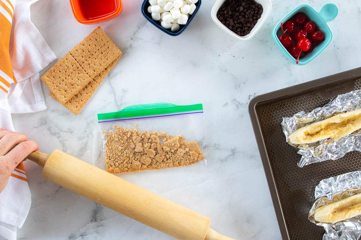 Graham cracker crumbs being crushed with a rolling pin with various ingredients surrounding the bag of crumbs.