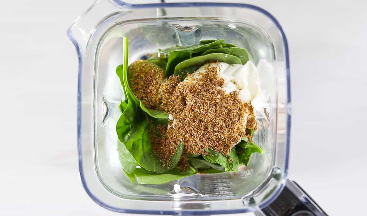 A top down view into a blender showing spinach, yogurt, and flax seeds.