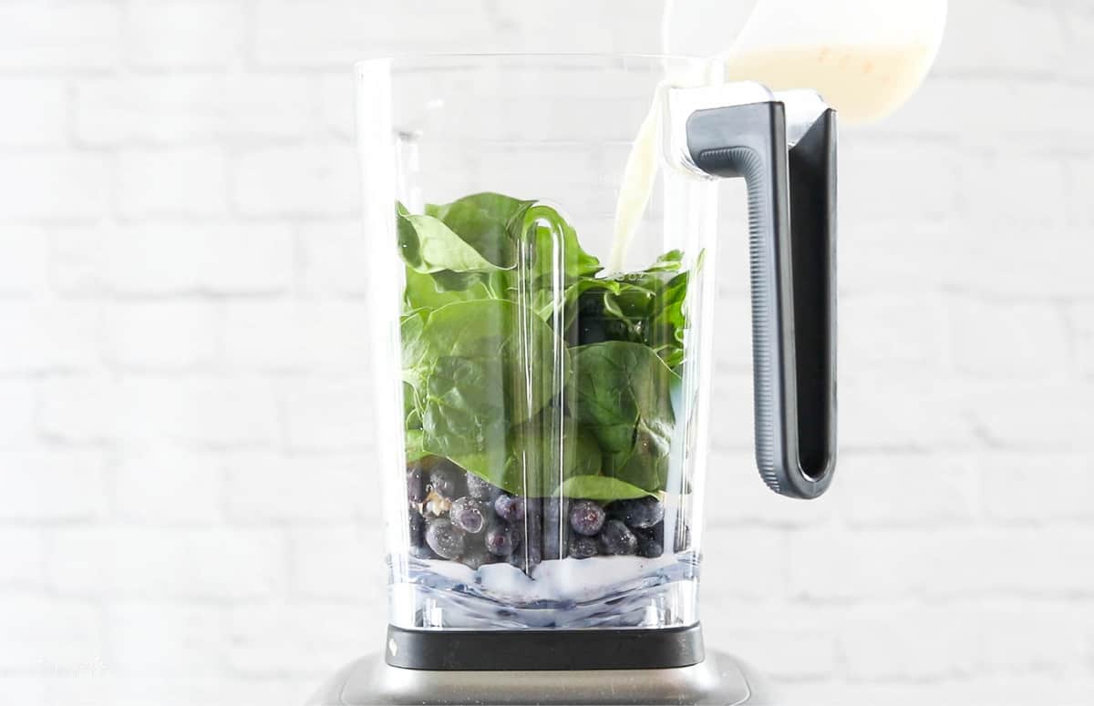 A front view of a blender with blueberries, spinach, and milk being added.