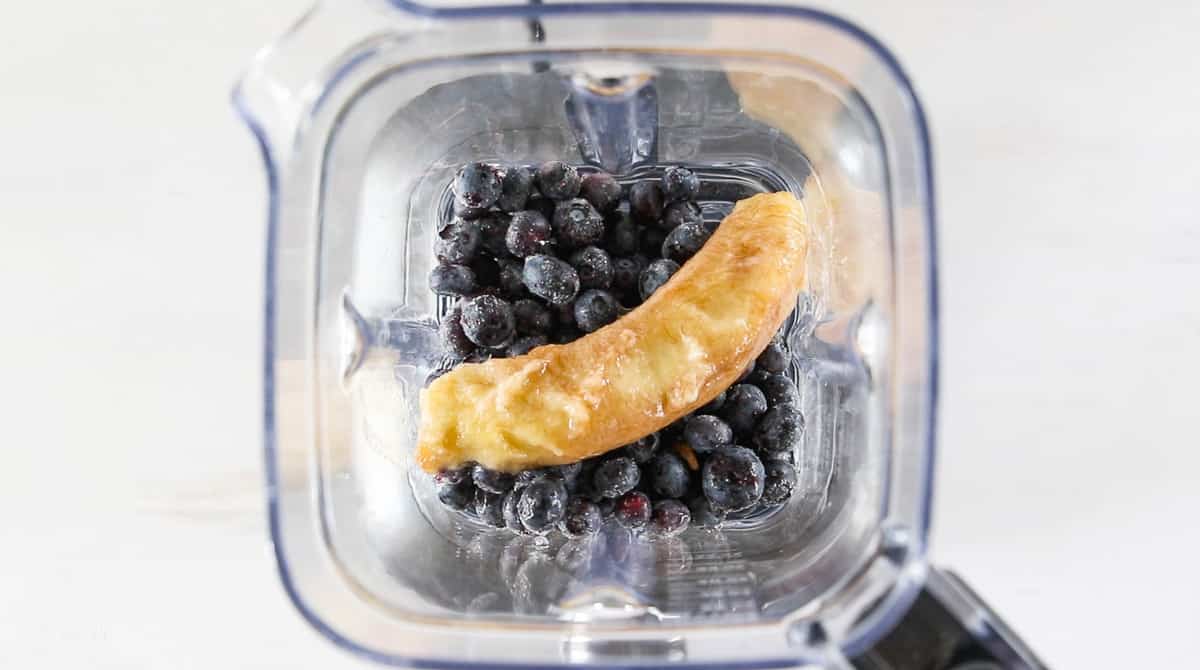 A top down view into a blender with frozen blueberries and a frozen banana.