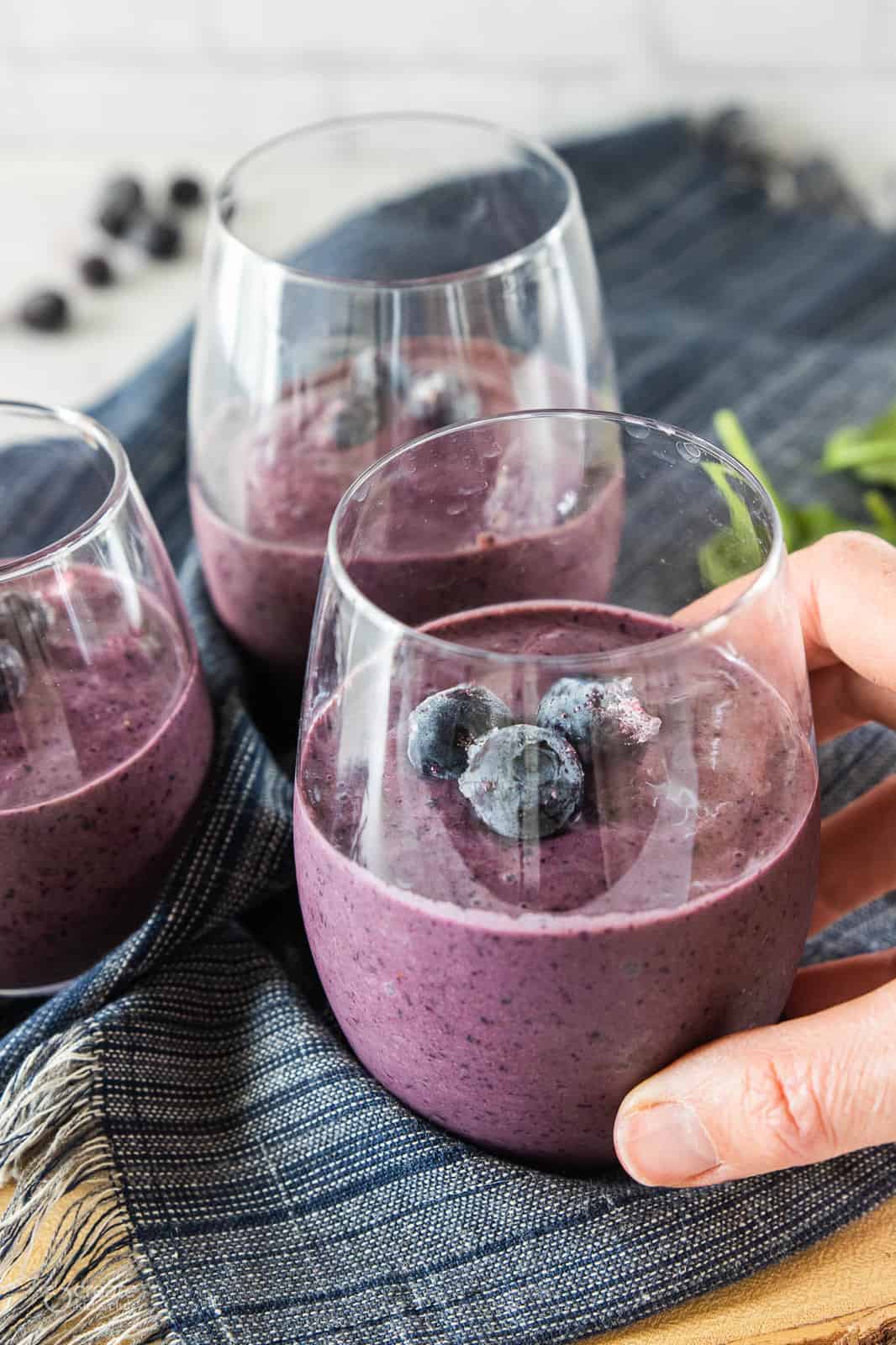 A high fiber blueberry spinach smoothie in a clear serving glass with a hand picking it up.