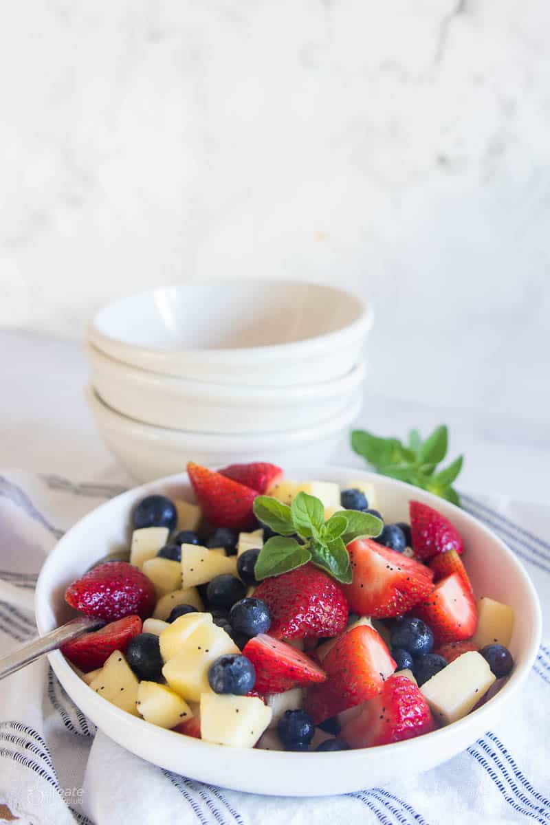 A summer salad with strawberries, blueberries, and apple in a white bowl.