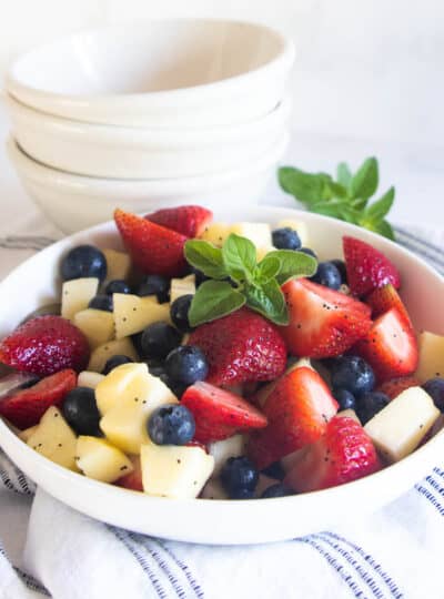 A summer salad with strawberries, blueberries, and apple in a white bowl with a poppy seed dressing.