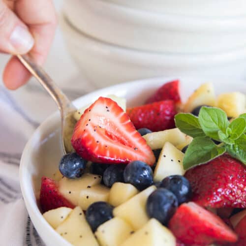 A hand spooning out a scoop of a fruit salad with strawberries, blueberries, and apple..