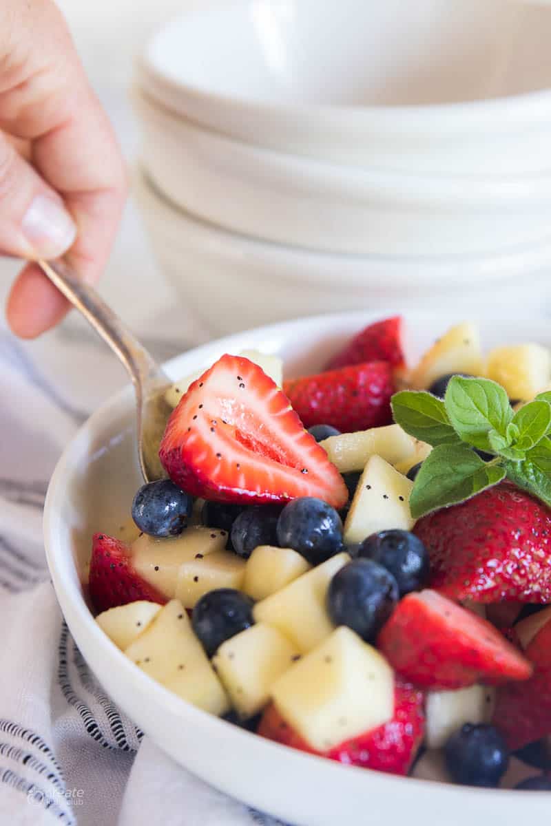 A hand spooning out a scoop of a fruit salad with strawberries, blueberries, and apple..