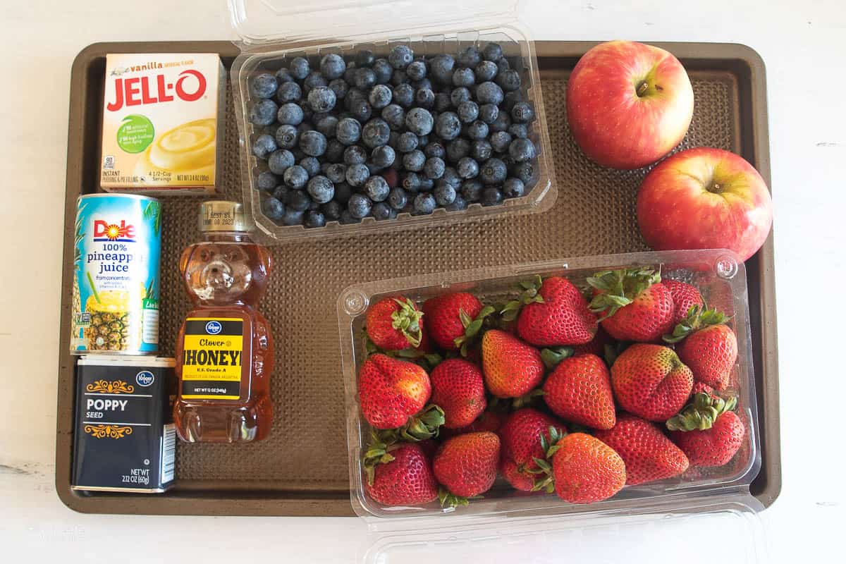 A tray with blueberries, strawberries, apples, jello mix, pineapple juice, honey, and poppy seeds.