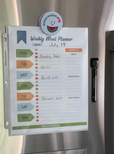 Weekly meal planner spreadsheet printed and placed in a sheet protector shown hanging on a refrigerator with a dry erase marker next to it.