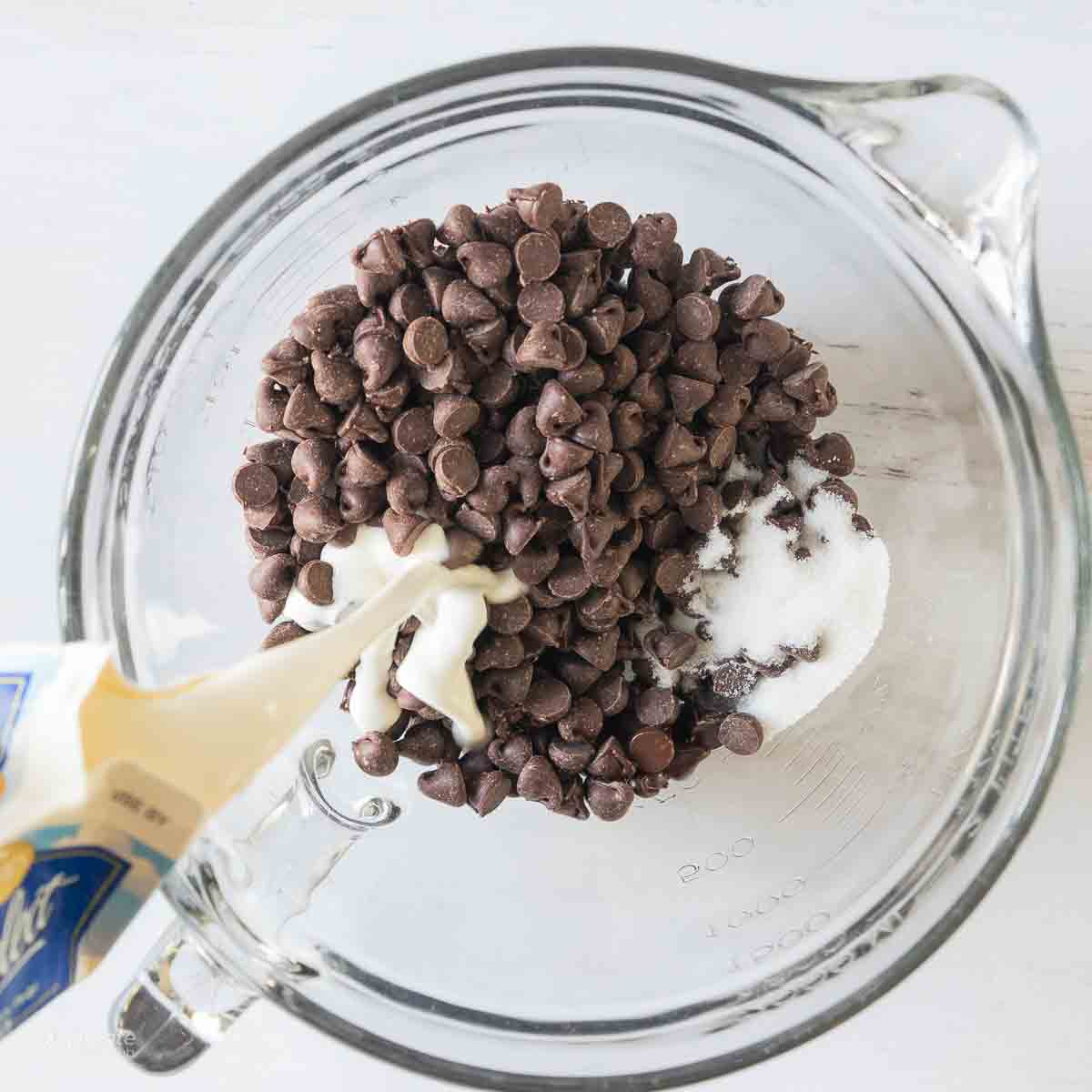 Chocolate chips in a glass bowl with sugar and whipping cream being poured in.
