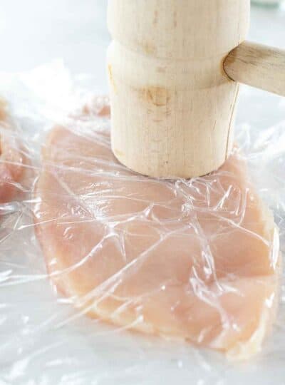 A thin chicken breast covered in plastic wrap with a mallet.
