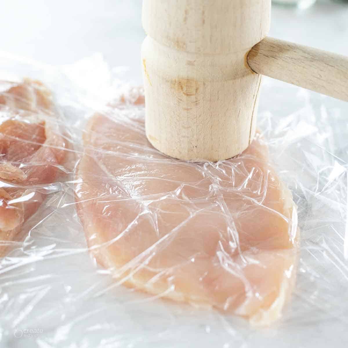 A thin chicken breast covered in plastic wrap with a mallet.