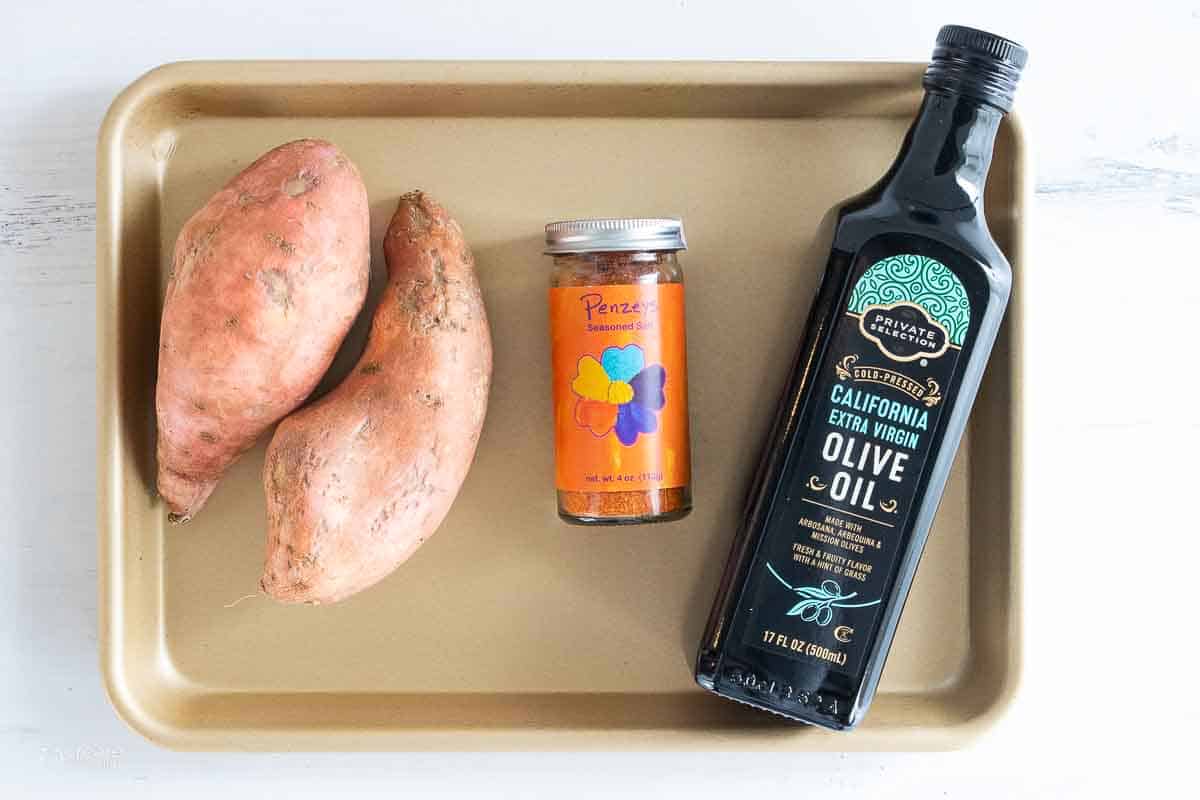 two sweet potatoes, jar of seasoning salt and bottle of olive oil on a sheet pan