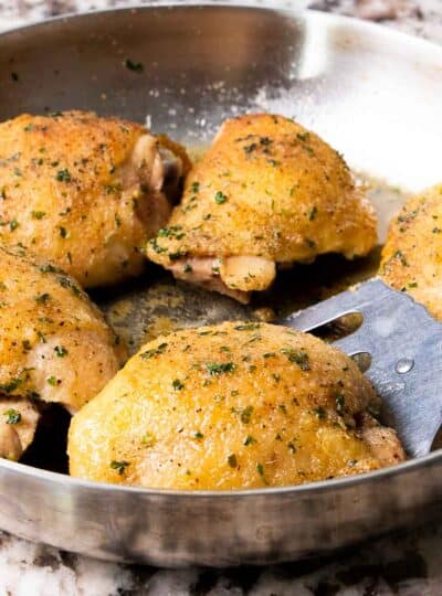 A close up of baked chicken thighs in a skillet with a spatula removing one.