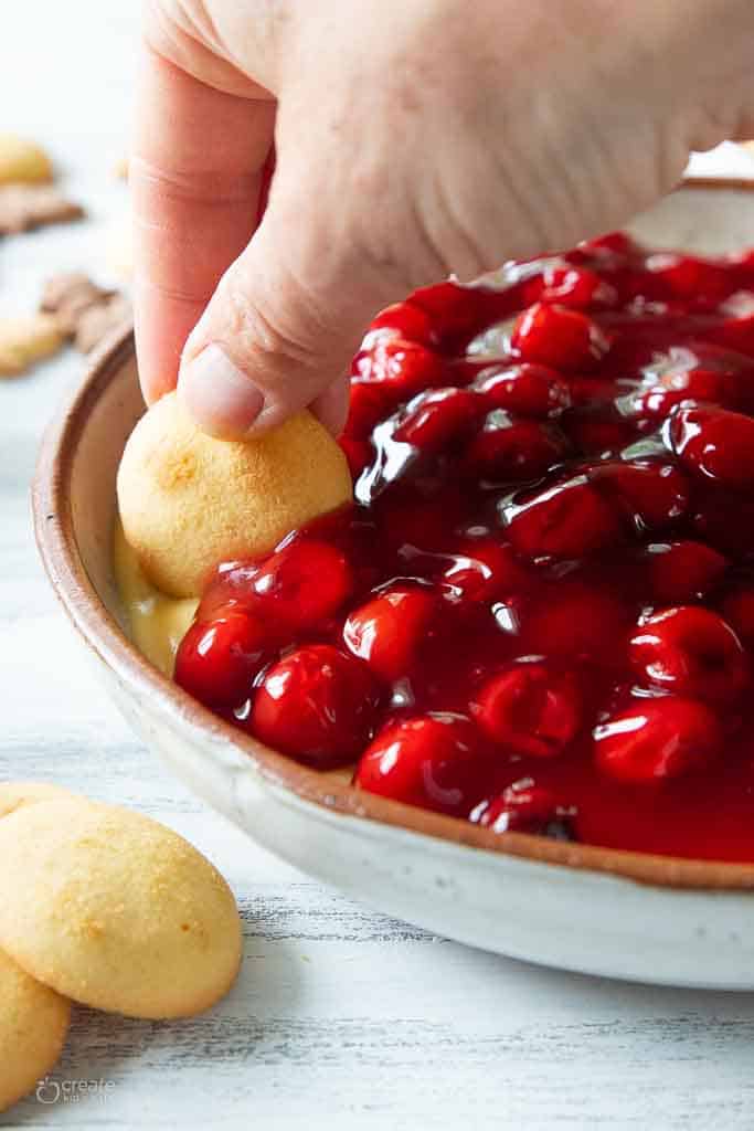 A hand dipping a vanilla wafer in cherry cheesecake dip.