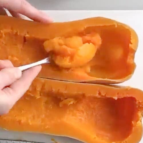 A hand using a spoon to scoop the insides from a cooked butternut squash sliced in half.