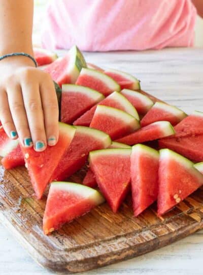 A little girls hand picking up a slice of watermelon off a wooden cutting board.