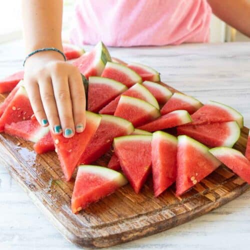 A little girls hand picking up a slice of watermelon off a wooden cutting board.
