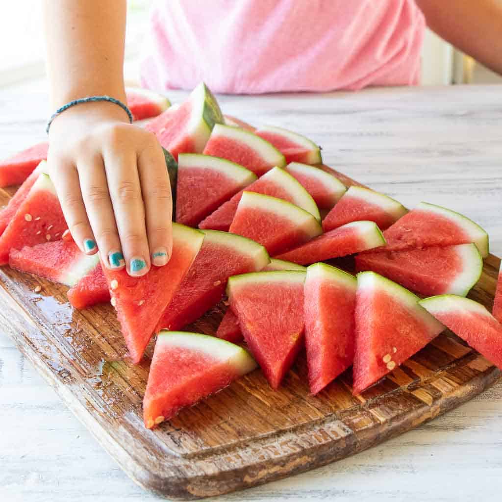 A little girls hand picking up a slice of watermelon off a wooden cutting board.
