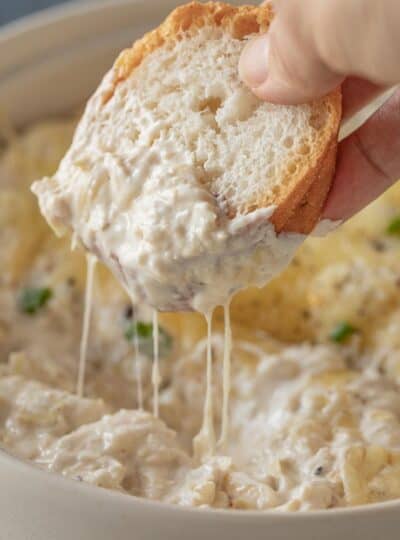 Crab artichoke dip showing a gluten free bread wedge scooping a cheesy bite.