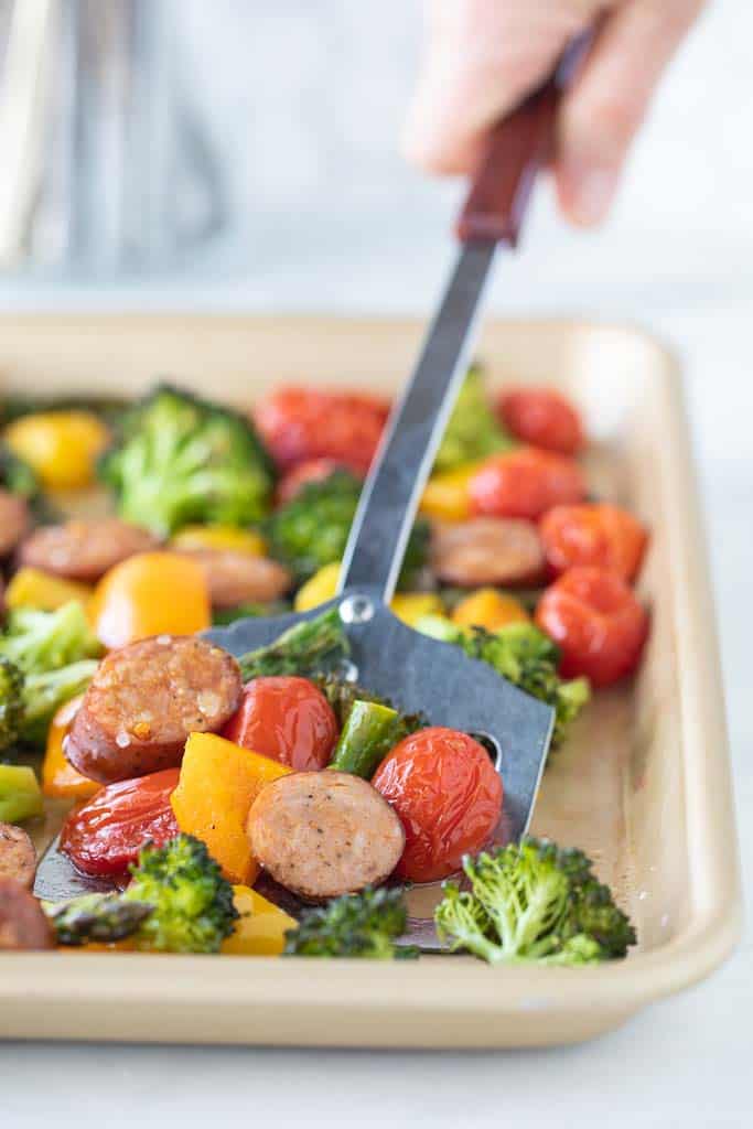 spatula scooping sausage and vegetables from sheet pan