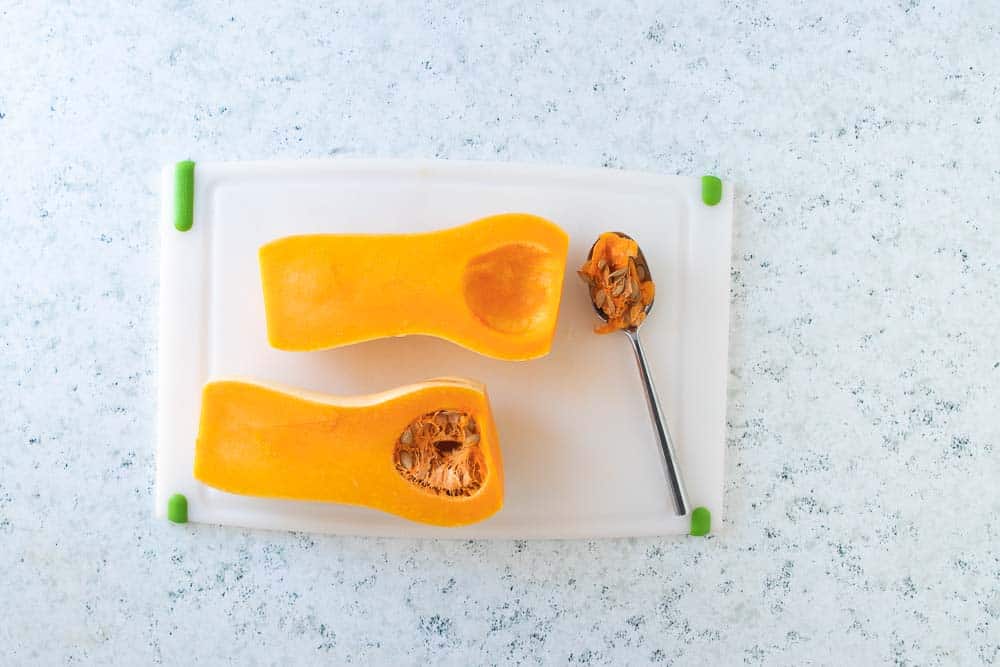A butternut squash sliced in half on a cutting board with a spoon scooping the seeds.