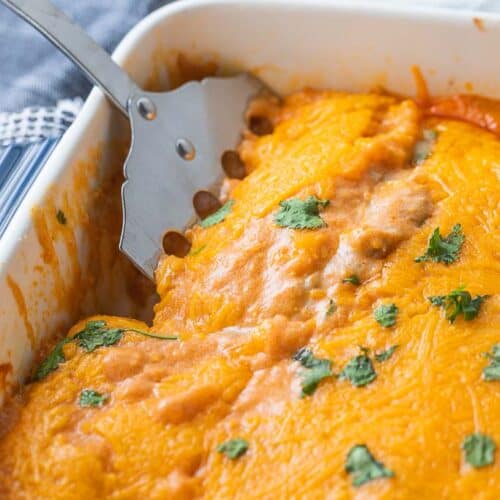 spatula in baking dish of enchiladas topped with melted cheese