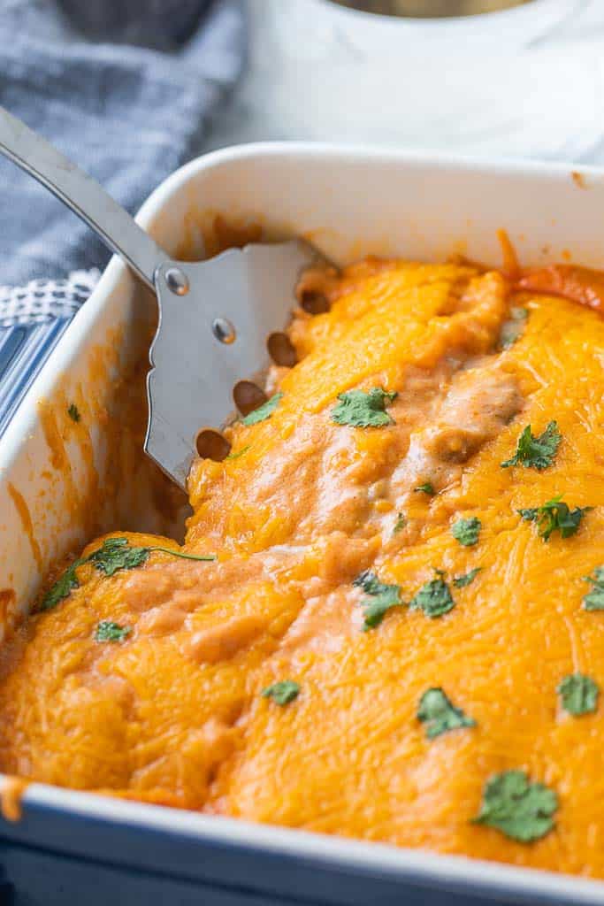 spatula in baking dish of enchiladas topped with melted cheese