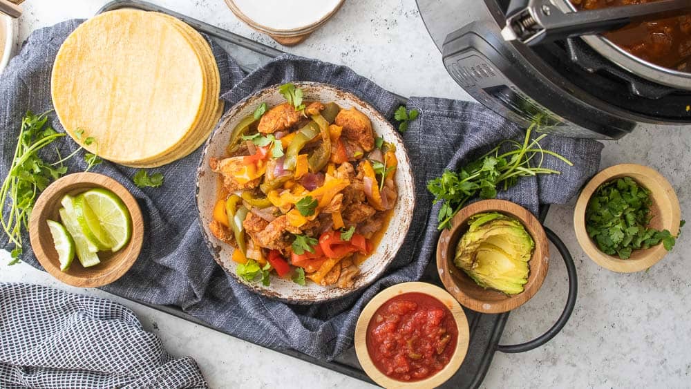 top down shot of bowls filled with lime wedges, salsa, avocado, cilantro surrounding large bowl of chicken fajitas