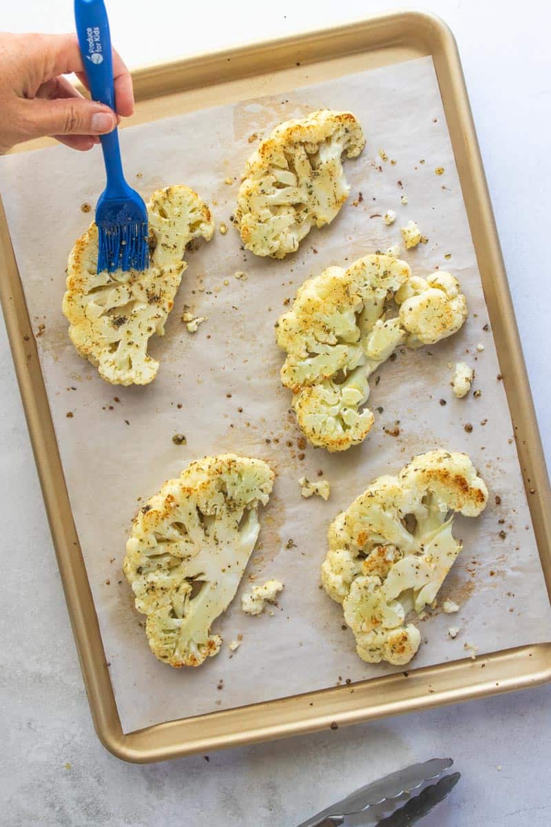 hand using pastry brush to cover cauliflower steaks with oil and spices